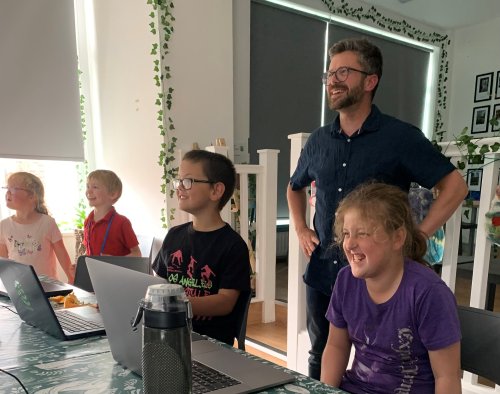 Children sitting at a table at The Bank, smiling with laptops, and a man standing with them smiling