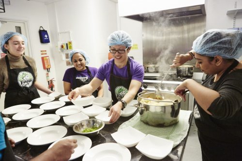 Shows food being dished up and lots of people gathered round a commercial kitchen table laden with plates.