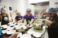 Shows food being dished up and lots of people gathered round a commercial kitchen table laden with plates.