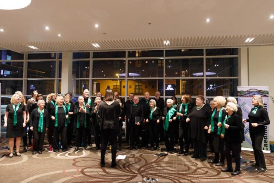 The choir rehearsing. The group are standing in a semi circle wearing black, with either green scarves or bow ties  