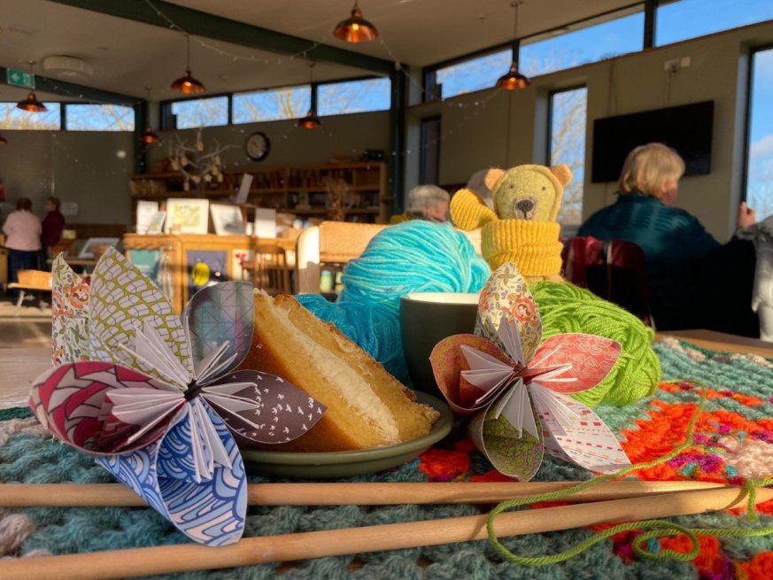 wooden knitting needles on a crochet blanket with a slice of cake and a cuppa, plus crafted flowers, cuddly toy and wool