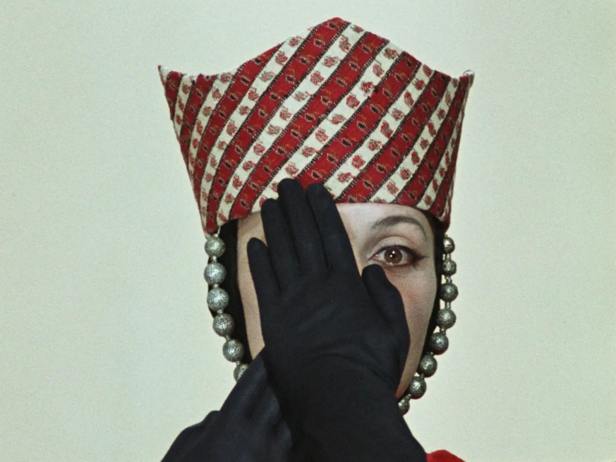 A film still showing a woman wearing a regal looking red striped hat with black gloves covering her face.