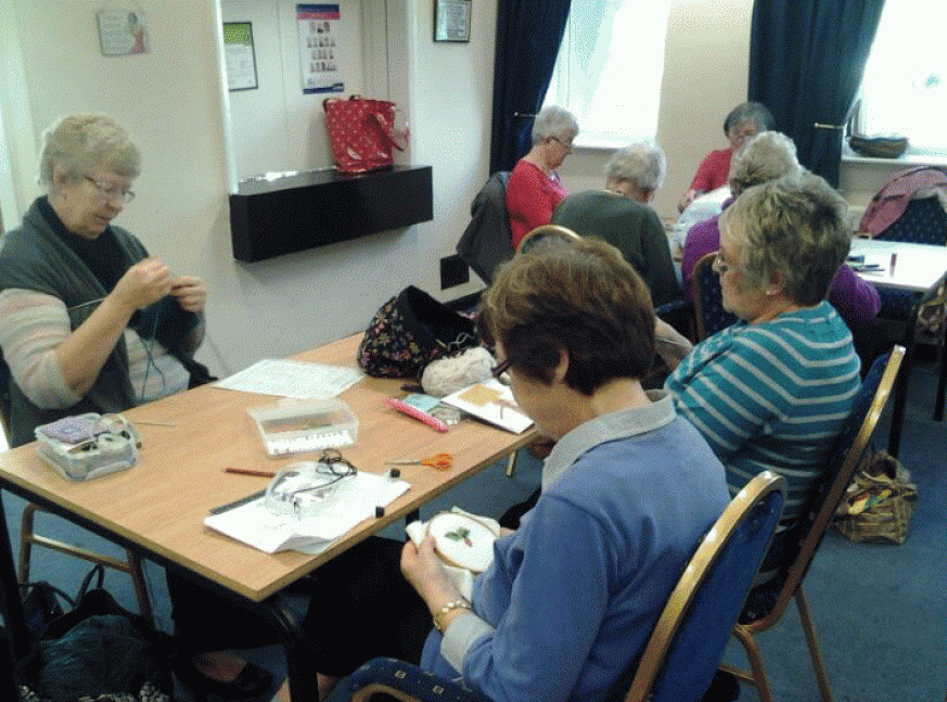 Members of the craft group sitting around a table busy working on knitting and embroidery 