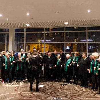 The choir rehearsing. The group are standing in a semi circle wearing black, with either green scarves or bow ties  