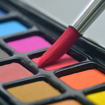 Photo of an art brush being dipped into red water colour paint with other colours also on view 