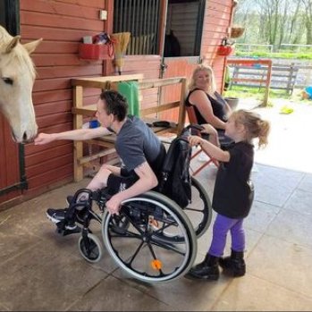 Photograph of a young man leaning froward from his wheelchair and allowing a white horse to smell his out reached fist. A much younger girl is holding the handles of the wheelchair . 