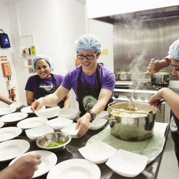 Shows food being dished up and lots of people gathered round a commercial kitchen table laden with plates.