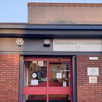 Front of clubhouse with sign.  red brick façade with grey framed glass door.